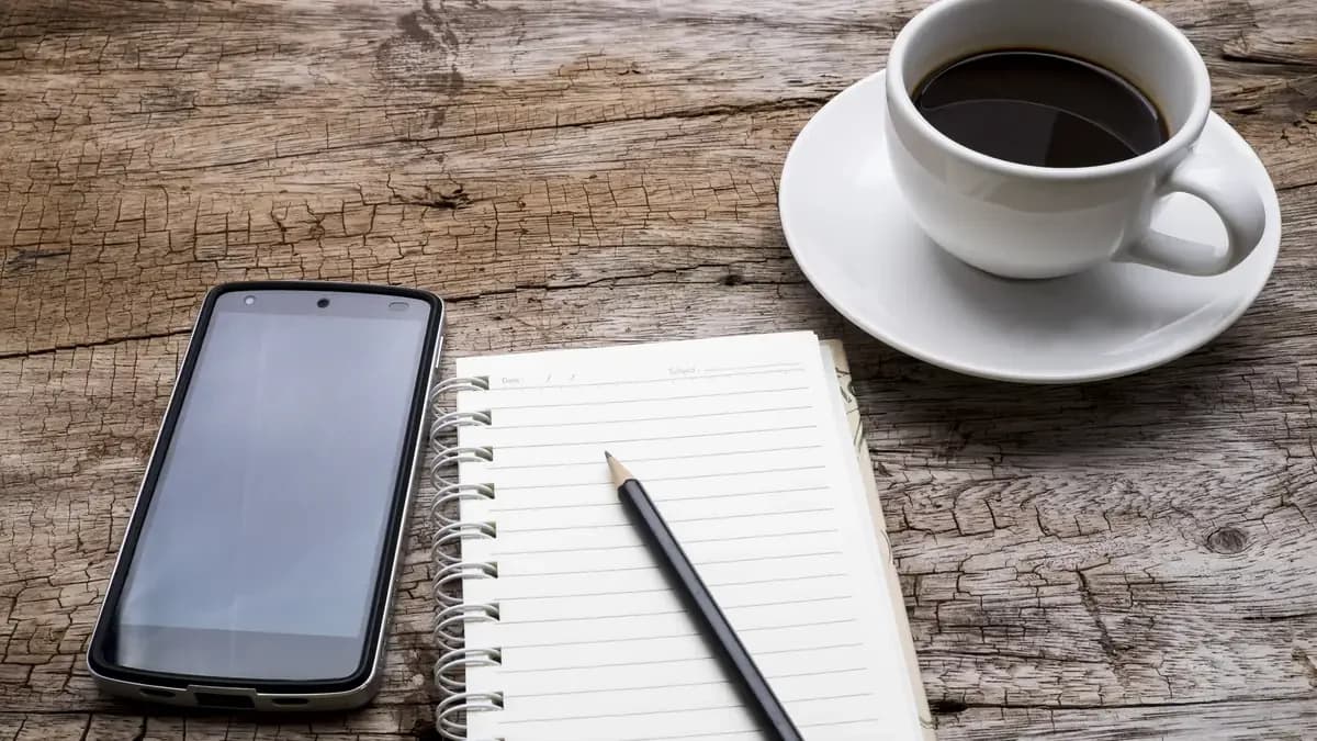 A spiral notebook with a pencil beside a phone and a cup of coffee on a wooden table