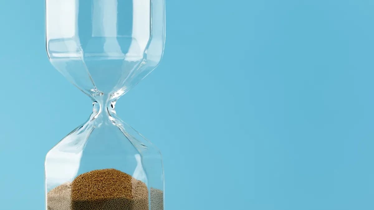 A clear glass sandglass with fine golden sand in the lower bulb against a plain blue background