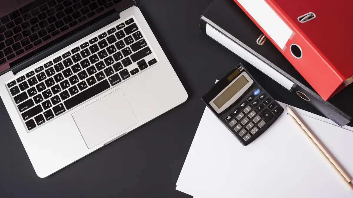 Laptop, calculator, blank paper and red folders laid out on a dark desk